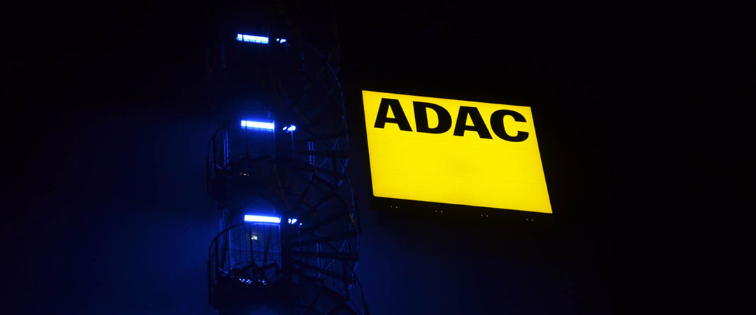 Gelb leuchtendes ADAC-Logo bei Nacht, links davon eine Wendeltreppe, die von blauer Beleuchtung erhellt wird.