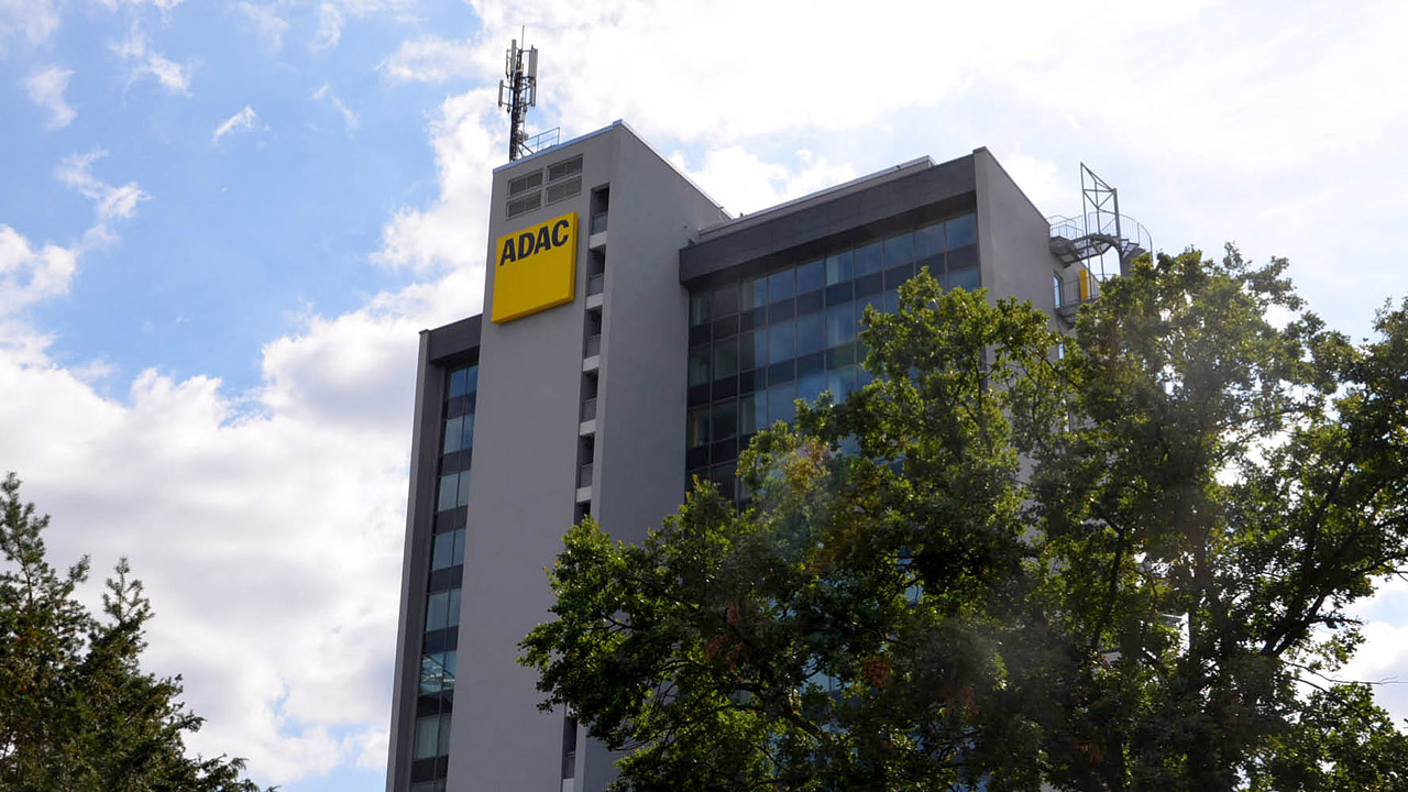 ADAC-Logo an einem hohen Bürogebäude in der Sonne mit Bäumen.