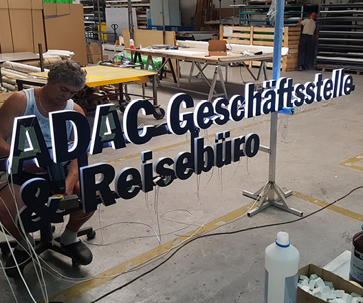 Ein Monteur ist in der Spiegelung der auf der Glasfläche angebrachten Leuchtschrift „ADAC Geschäftsstelle & Reisebüro“ zu sehen.