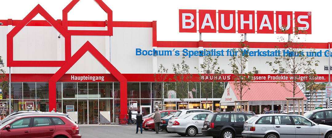 Eingangsbereich eines BAUHAUS-Marktes mit roter Fassadengestaltung, mehreren geparkten Autos und der Beschriftung „Bochum's Spezialist für Werkstatt, Haus und Garten“.