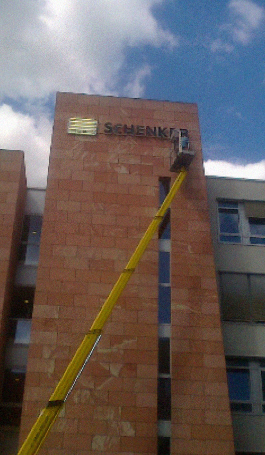 Arbeiter installiert DB Schenker-Leuchtschrift auf einer gemauerten Fassade eines Firmengebäude mithilfe eines Hubwagens.