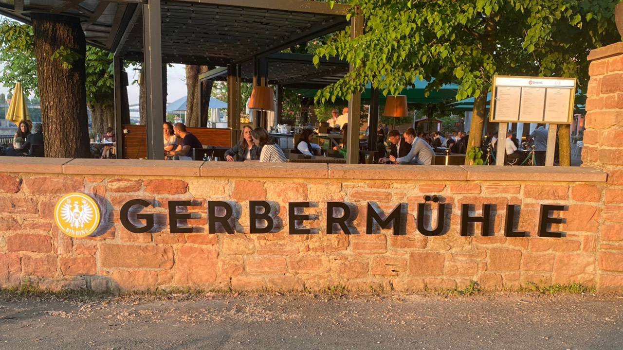 Gerbermühle Schriftzug auf einer Steinmauer in Frankfurt, umgeben von grünen Bäumen und mit Restaurantgästen im Hintergrund