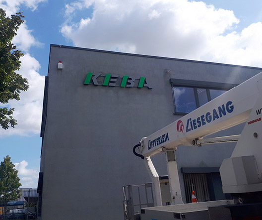 KEBA-Logo an einer hohen Betonwand, Montagearbeiten mit Hebebühne im Vordergrund.