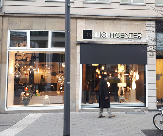 Außenansicht des LichtCenter-Geschäfts mit großen Fenstern und Designerleuchten im Schaufenster sowie schwebend installiertem LichtCenter-Logo.