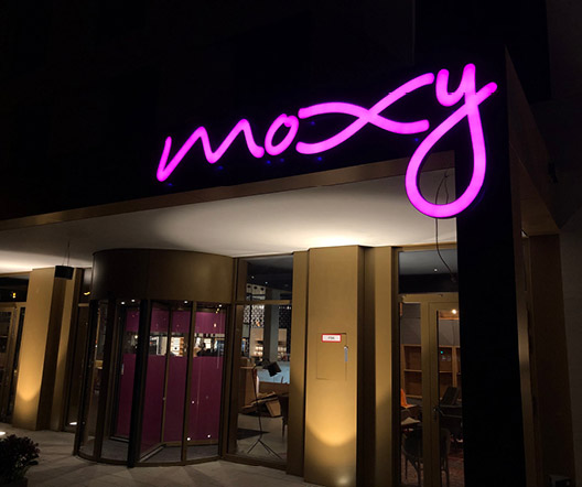 Eingang des Moxy Hotels in Darmstadt bei Nacht mit darüber angebrachter pinkfarbener Leuchtschrift