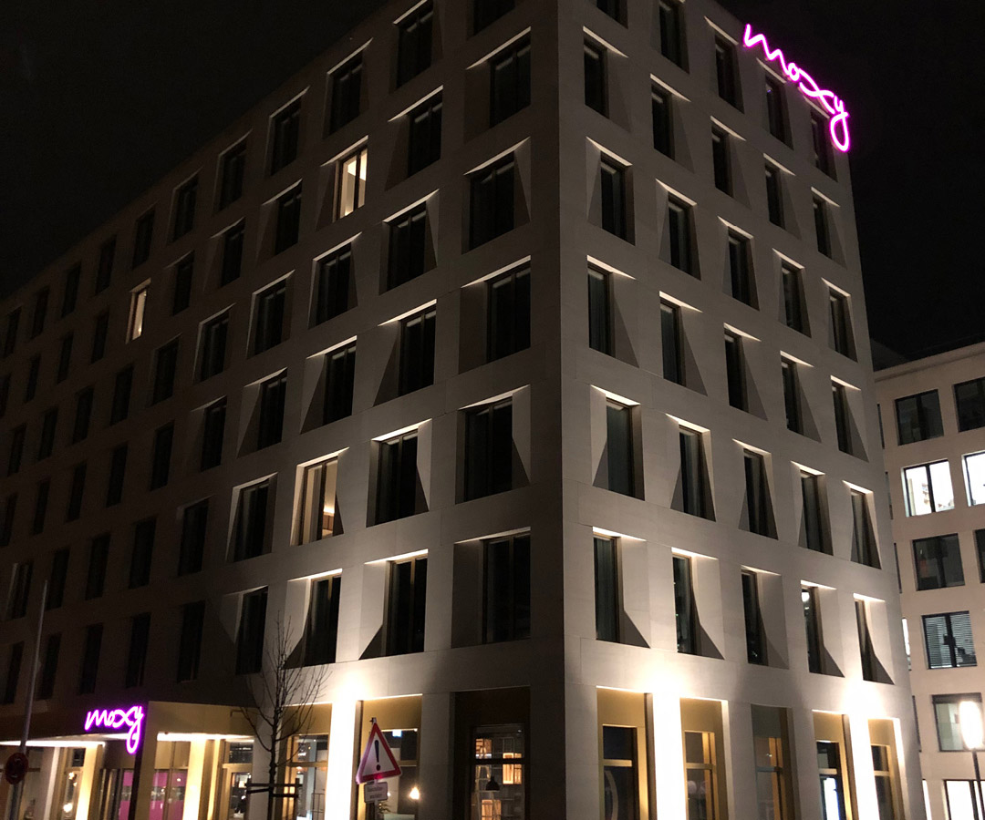 Moxy Hotels Gebäudefront bei Nacht mit beleuchteter Leuchtschrift in auffälligem Magenta.