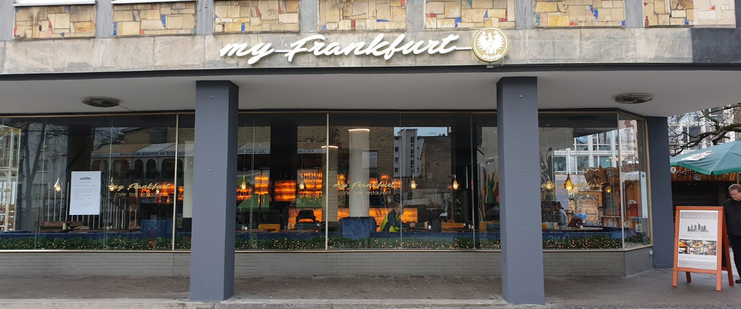 Frontalansicht des „My Frankfurt“ Cafés mit großen Fenstern, die den Innenraum sichtbar machen.