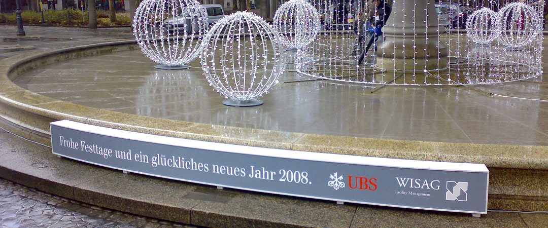 Seitenansicht des beleuchteten Brunnens vor der Alten Oper in Frankfurt mit einem Neujahrsgruß auf Deutsch von UBS und WISAG.