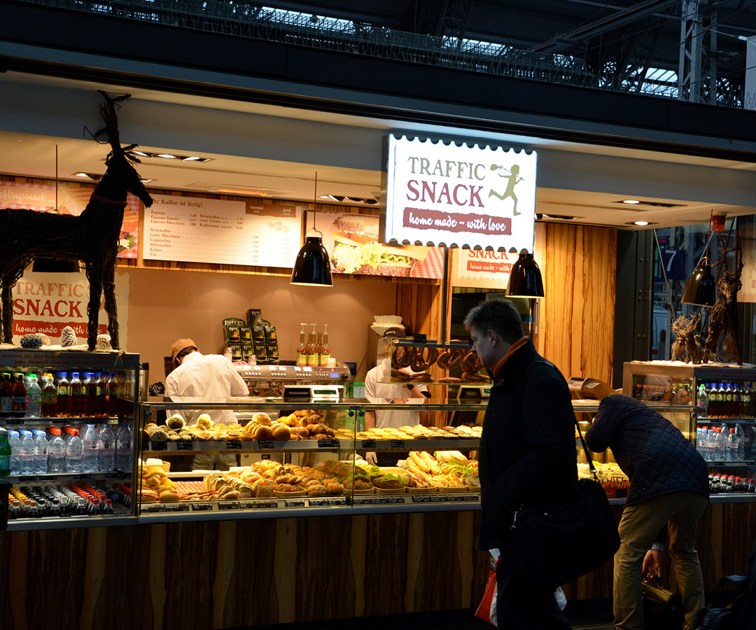 Verkaufsstand von „Traffic Snack“ mit beleuchtetem Schild und vielfältigem Angebot an Backwaren, Kunden stehen vor der Theke.